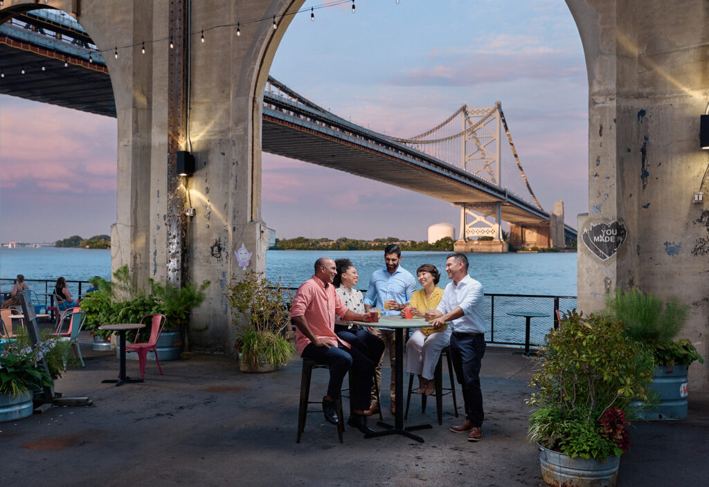 A group of people laugh at a table at Cheery Street Pier. The Ben Franklin Bridge is seen in the background.