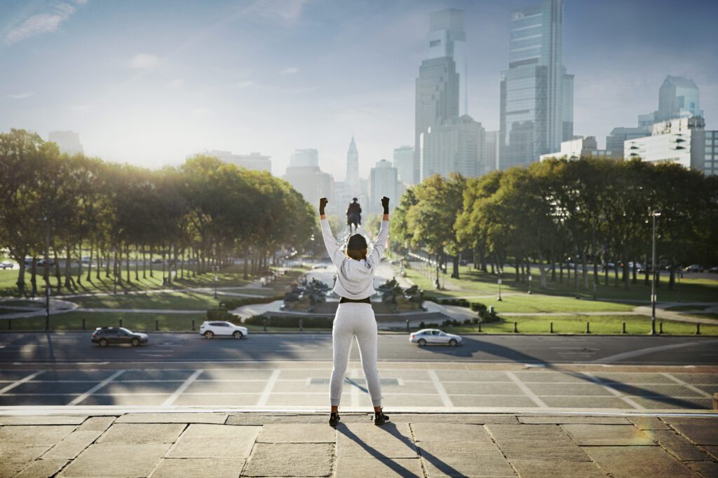 A woman stands on top of the Rocky steps, facing the Philadelphia skyline, with her arms raised above her head