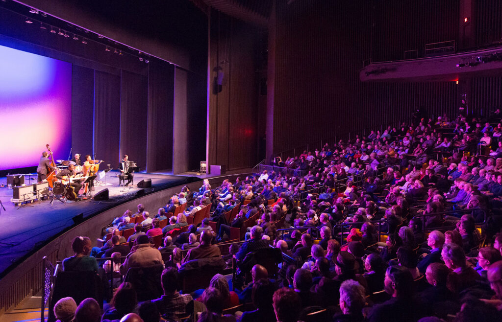A performance takes place on stage in front of a crowded theater.