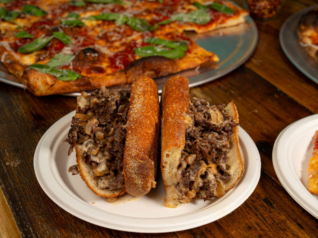 A cheesteak with a pizza behind it