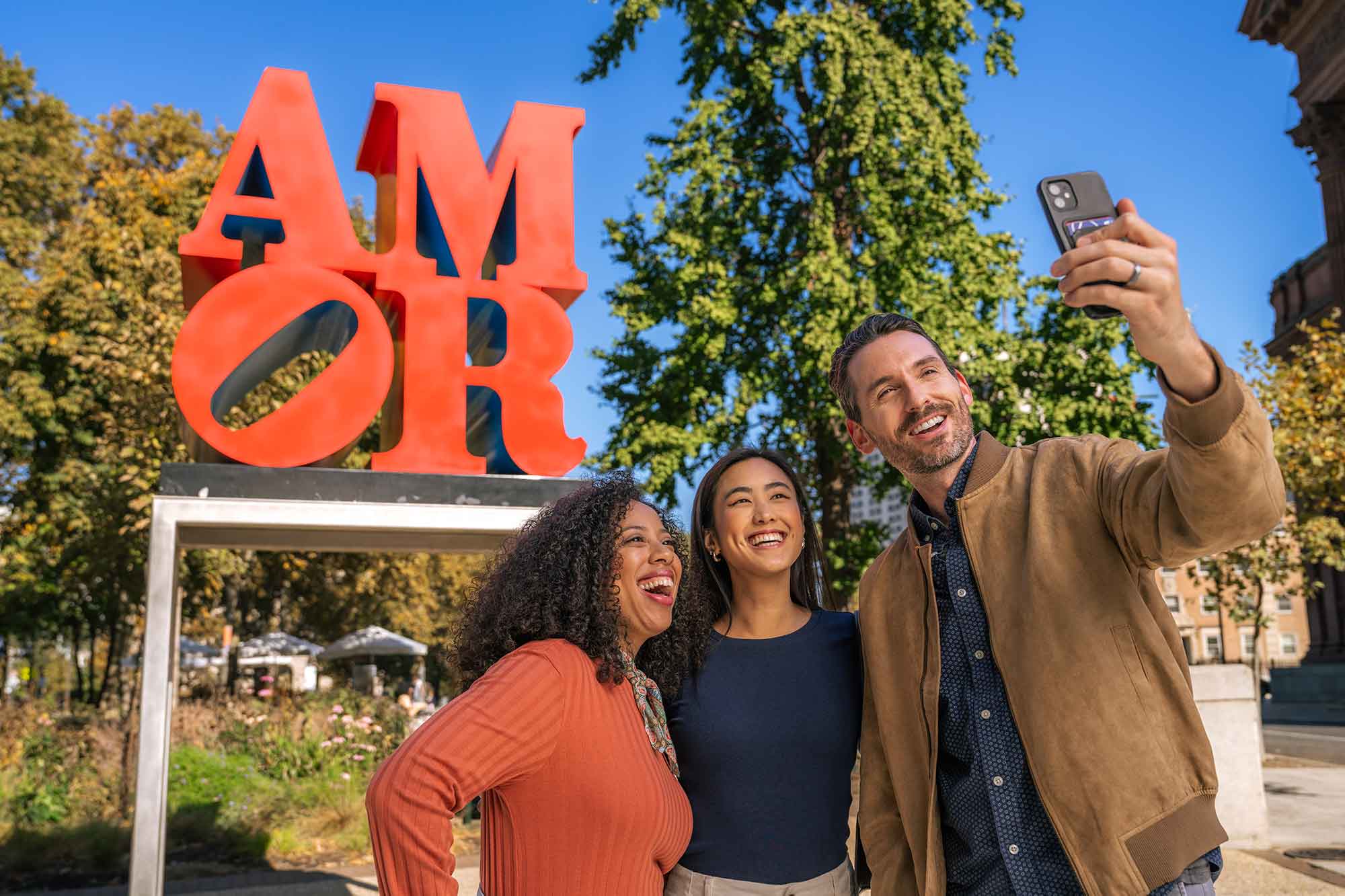 Three people pose for a selfie in front of the AMOR statue on Ben Franklin Parkway.