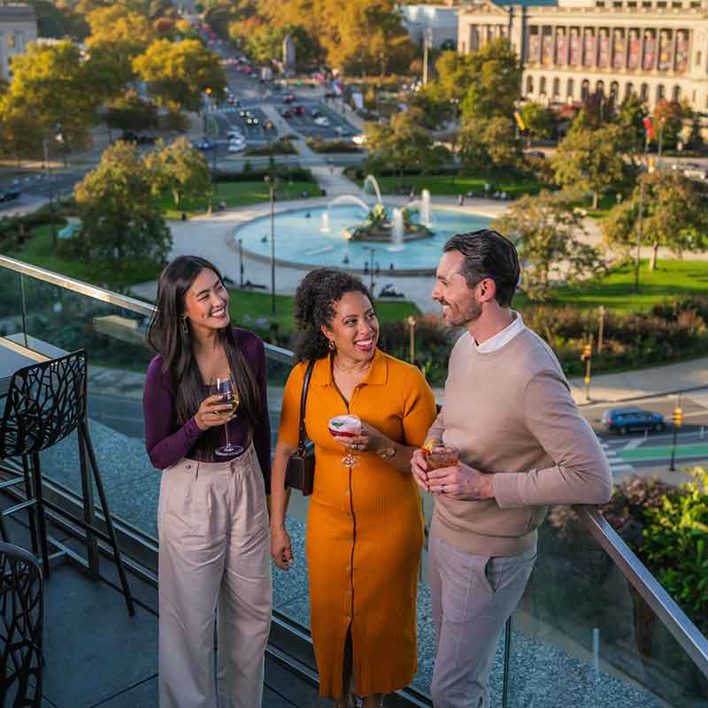 Three people laugh and talk on a rooftop overlooking Ben Franklin Parkway.