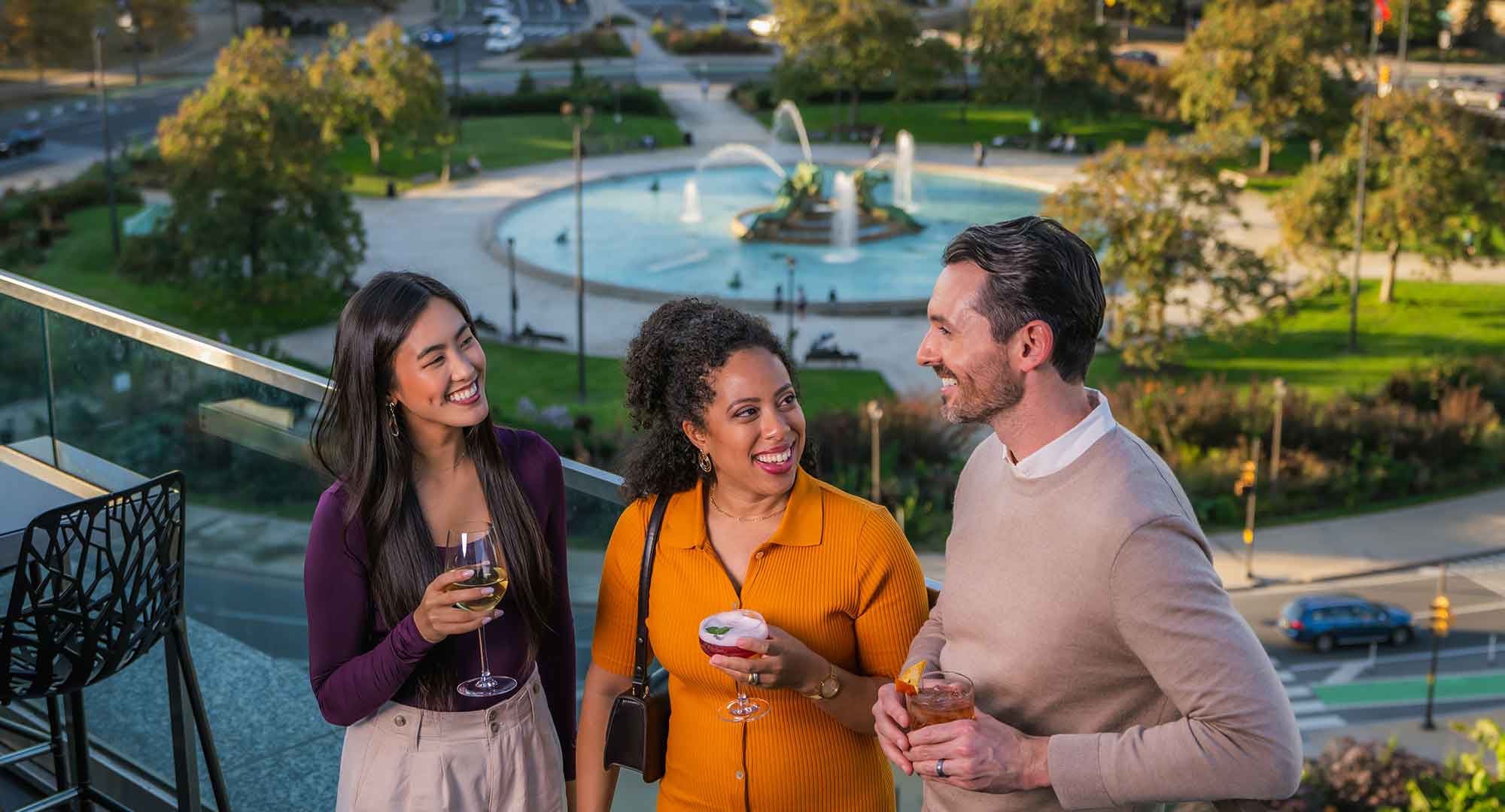 Three people laugh and talk on a rooftop overlooking Ben Franklin Parkway.