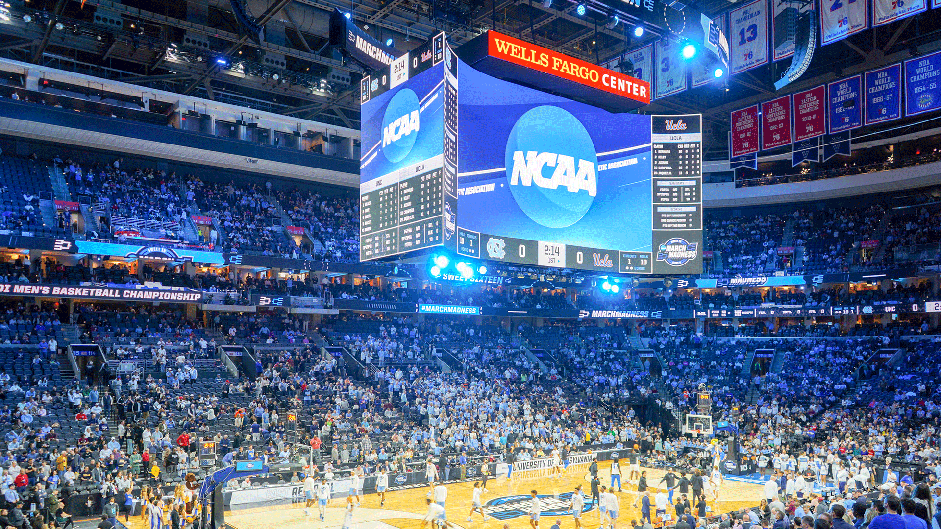 a basketball court is shown full of players. Coaches are lined up along the sides of the court. Fans are in the seats surrounding them. The jumbo tron hanging above the basketball court reads March Madness.
