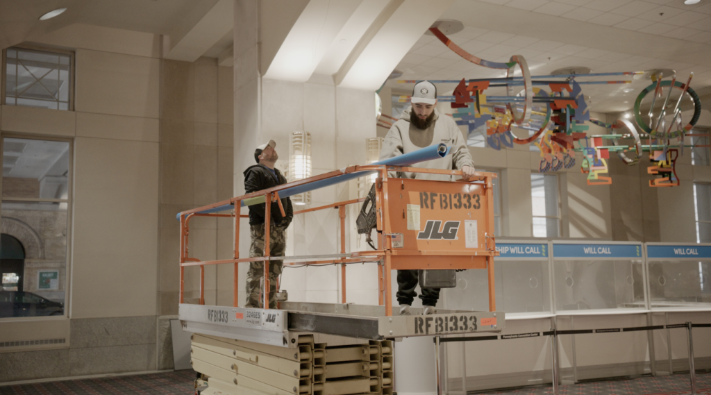 Two men stand on a lift, installing a sign in the PA Convention Center.