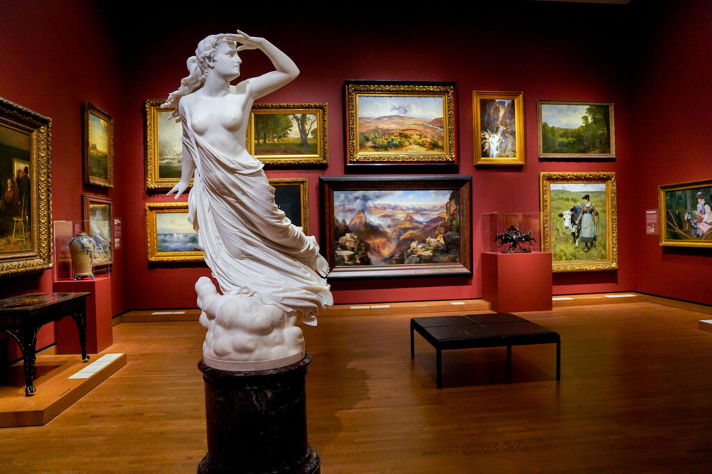 A marble statue sits in a deep red room, full of paintings on the walls.