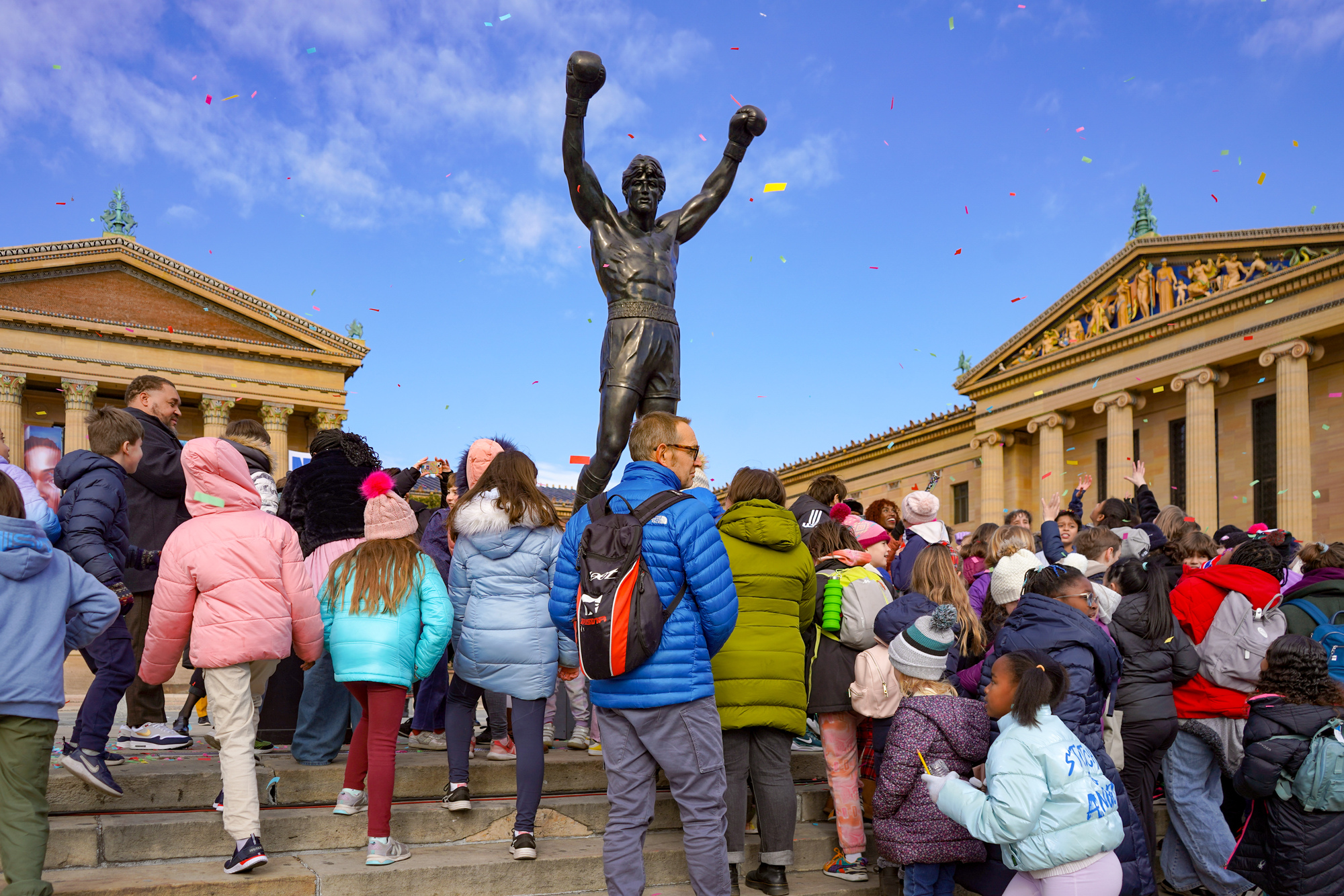 Many people and children at the top of the Rocky Steps at the Philadelphia Museum of Art. The Rocky statue is at the top of the steps and there is confetti from a celebration.