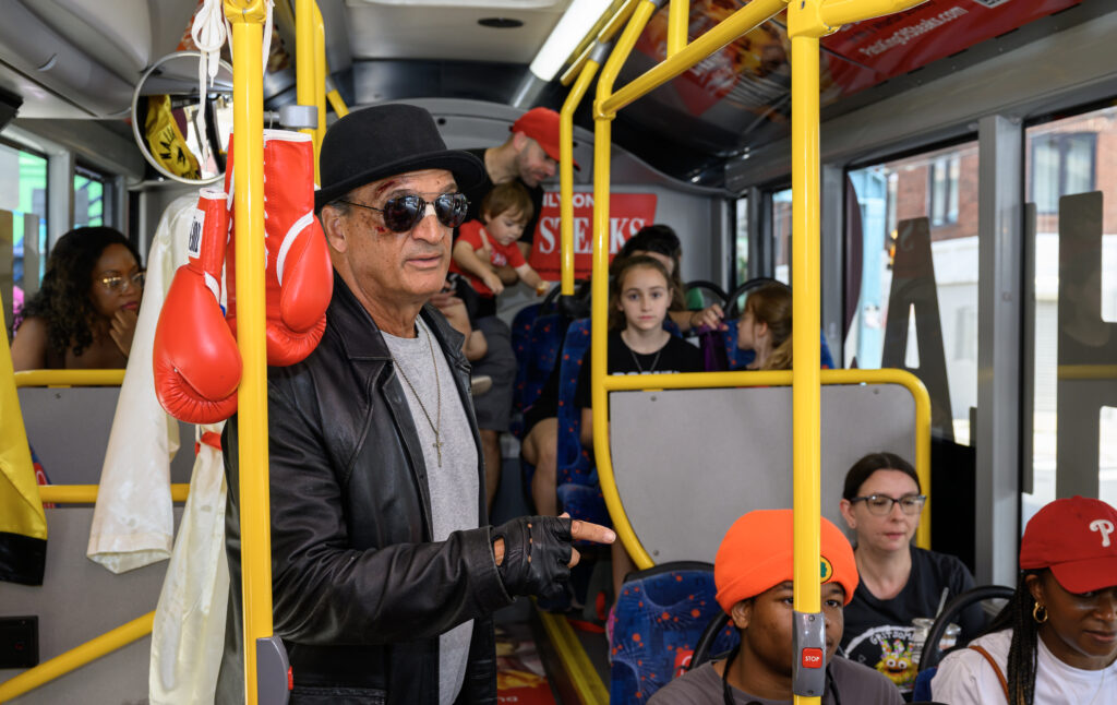 People on a bus with a Rocky impersonator in the foreground